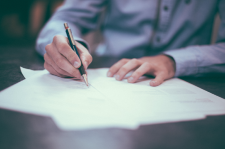 A man's hands hold a pen and write on documents laid across a wooden desk.